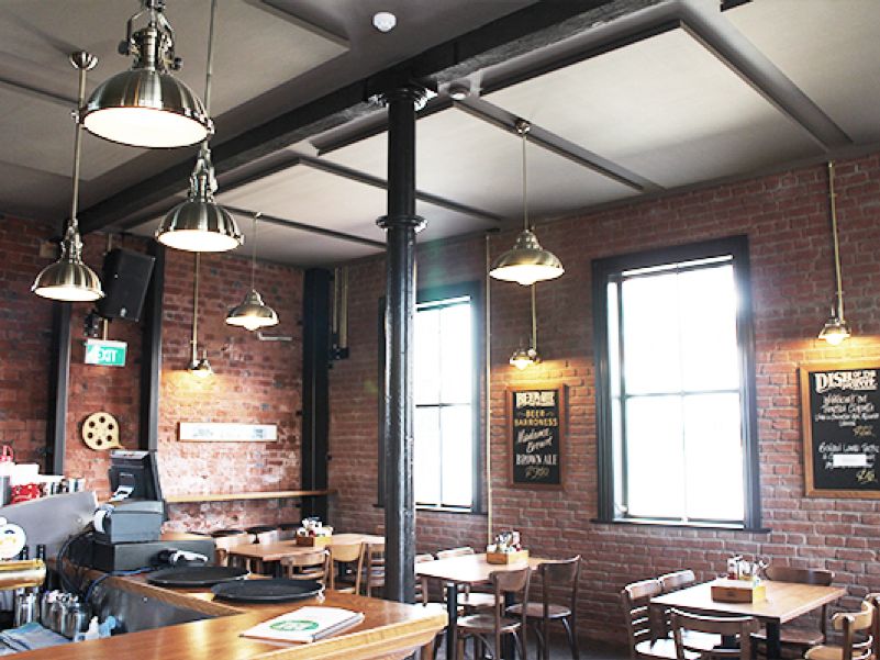 Industrial design restaurant, with wooden bar in the foreground, tables in the background, bare brick walls and light grey acoustic panels suspended from the ceilling