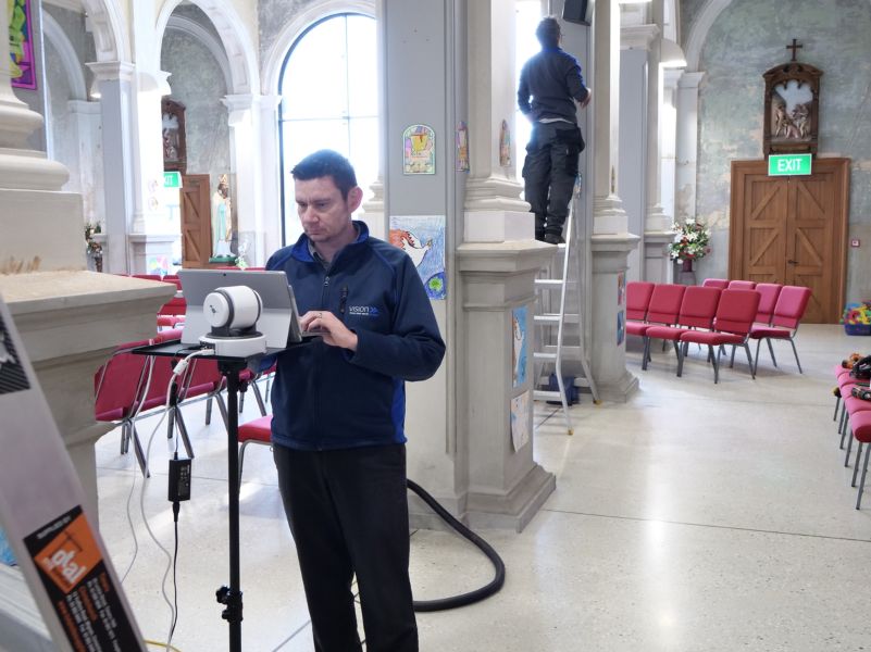 Neville on a laptop in the foreground, Andrew on a ladder in the background, inside St. Patrick's Basilica